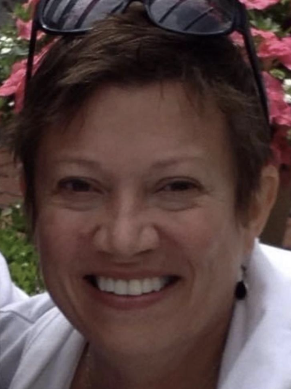 A person with short brown hair and sunglasses resting on their head smiles at the camera. Pink flowers and greenery are visible in the blurred background.